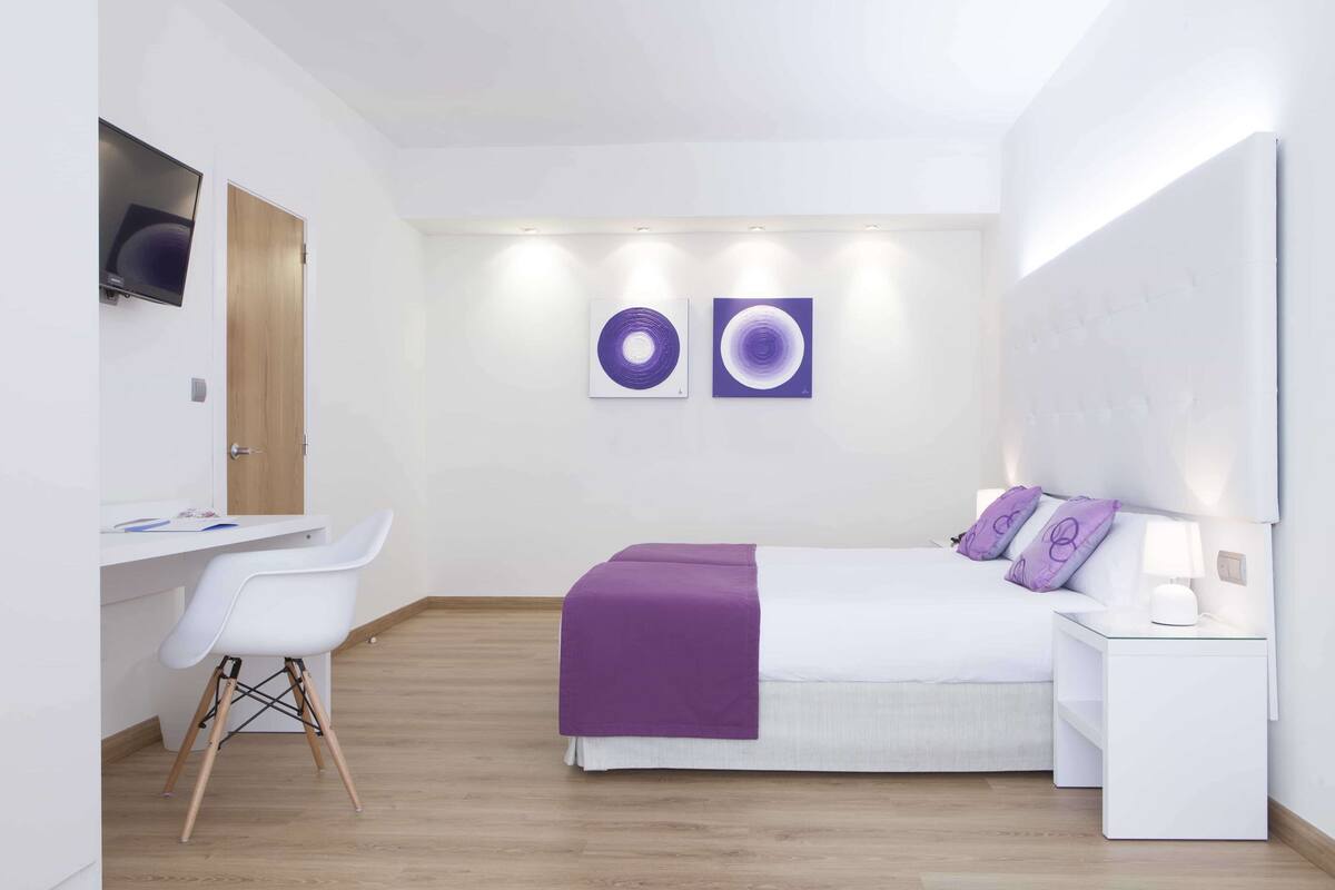 A modern hotel room with a bed, a desk, and a chair. There are two wall-mounted artworks and a flat-screen TV.