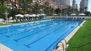 A swimming pool with lounge chairs and umbrellas, surrounded by palm trees and high-rise buildings. - Hotel Albahia (Alicante)