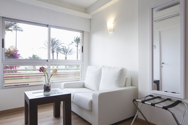 Una sala moderna con un sofá blanco, una pequeña mesa de madera con un jarrón y vistas a palmeras a través de amplios ventanales.