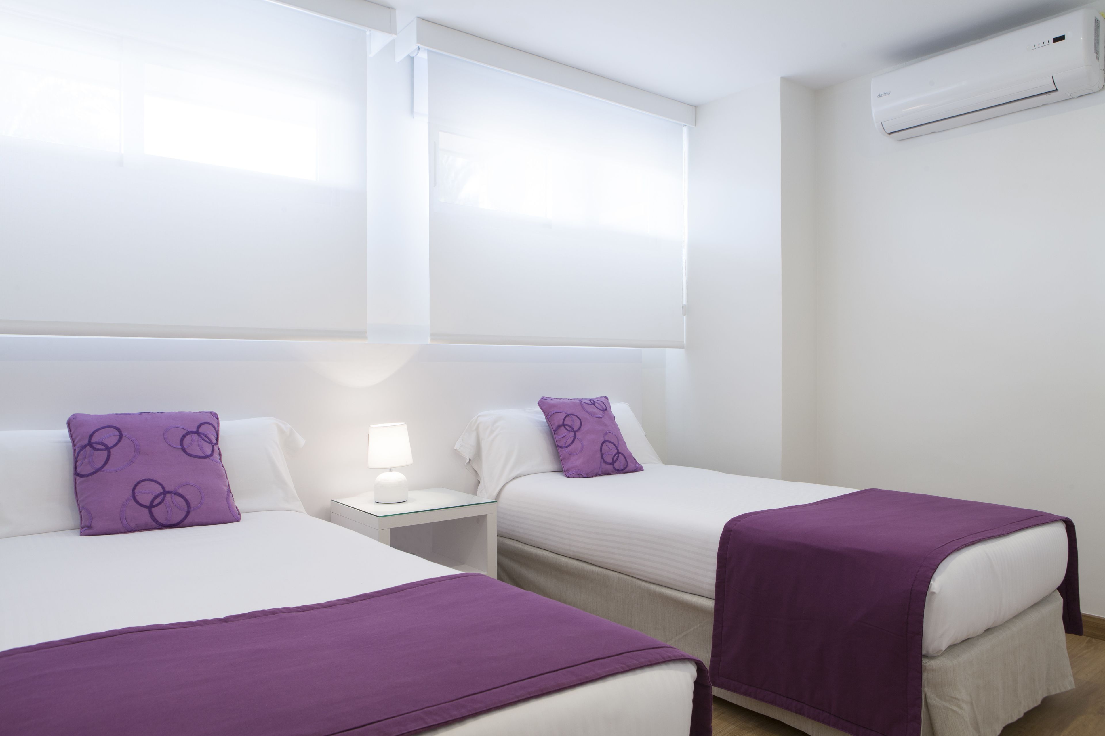 Une chambre d’hôtel avec deux lits, chacun recouvert d’une couverture et doté d’un oreiller violets, une table de chevet avec une lampe et un appareil de climatisation fixé au mur.
