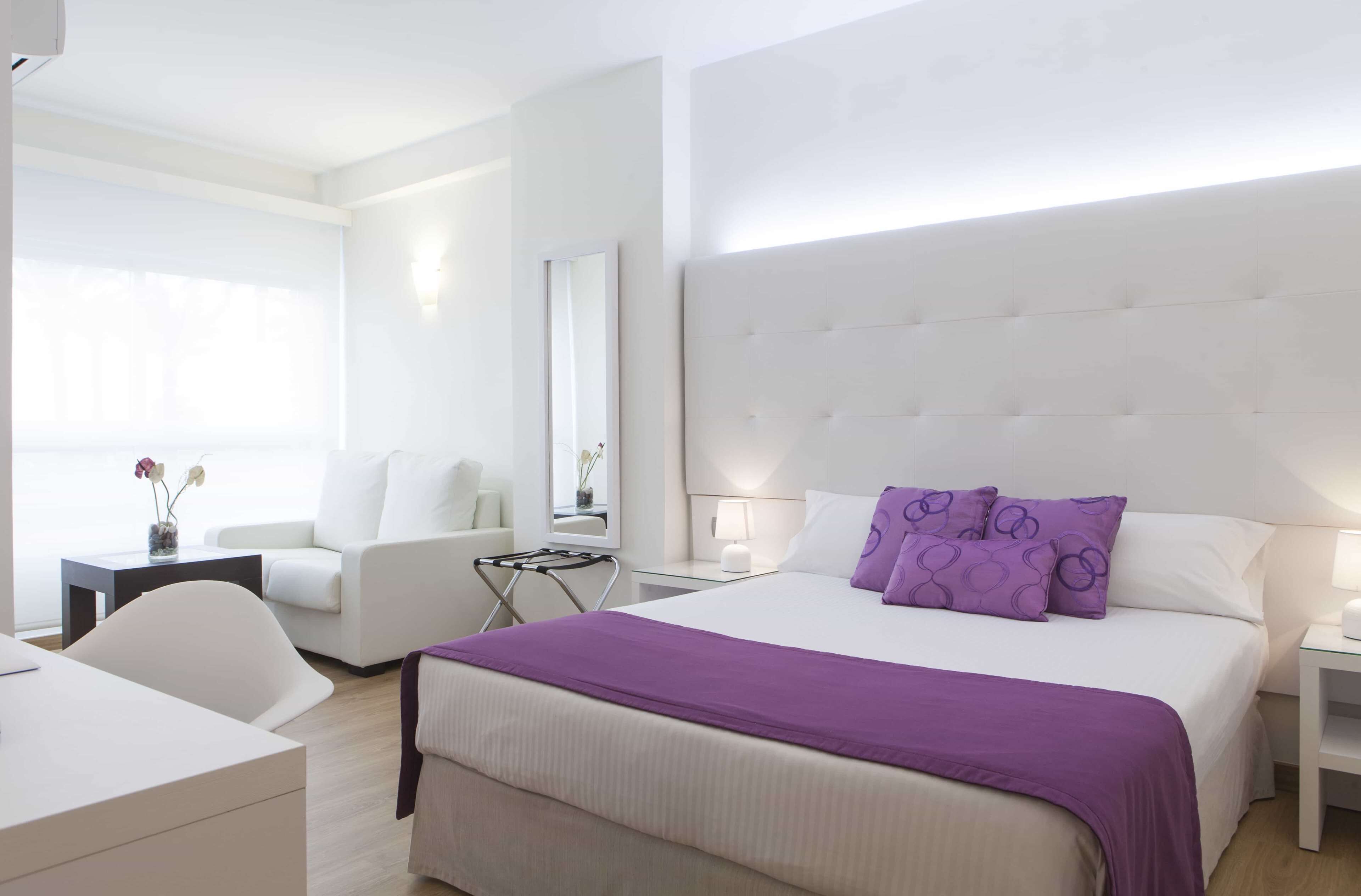 Una camera d'albergo moderna con un letto grande, una poltrona bianca, una scrivania e un comodino con una lampada.