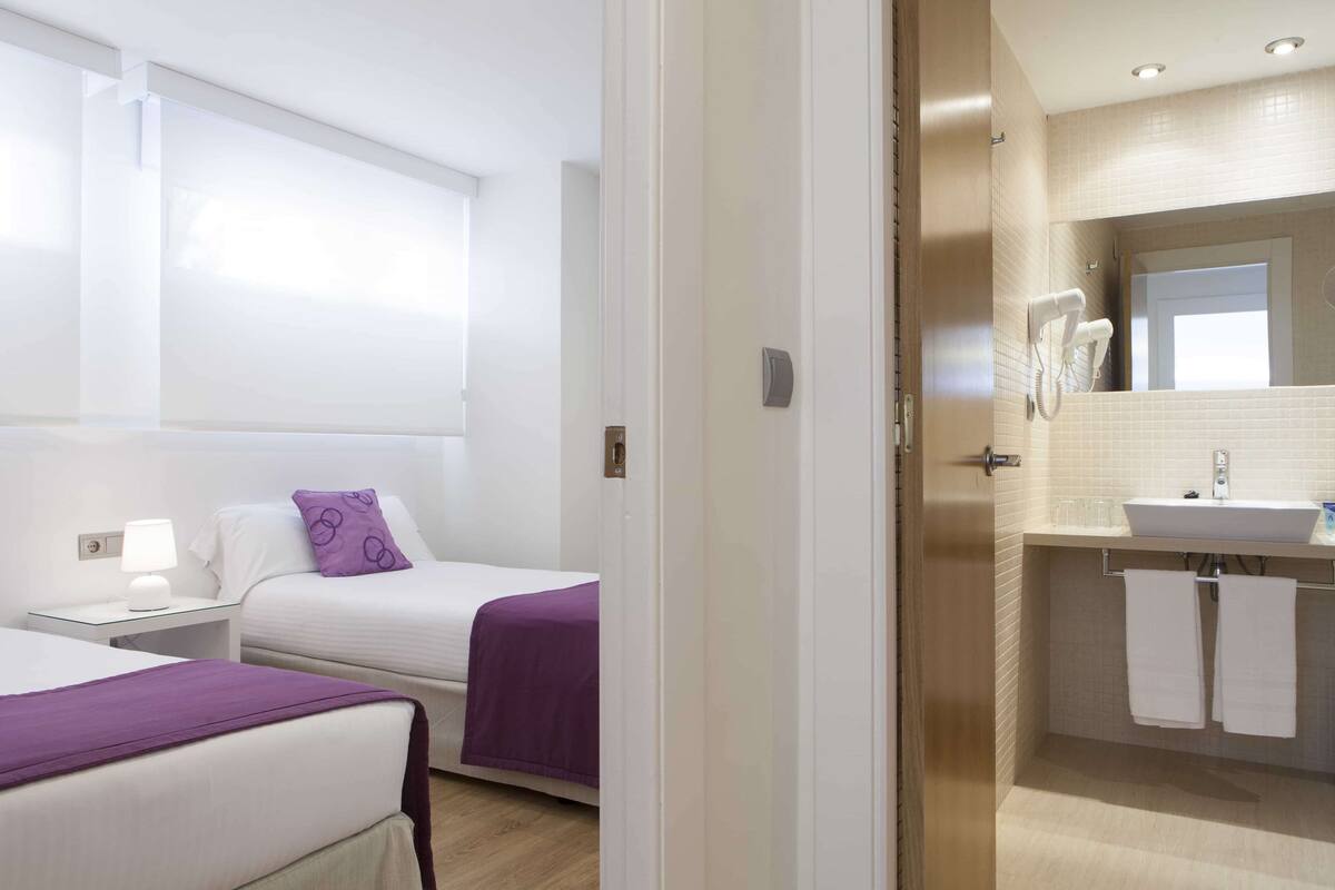 A hotel room with two beds, a bathroom with a sink and mirror, and a window with blinds.