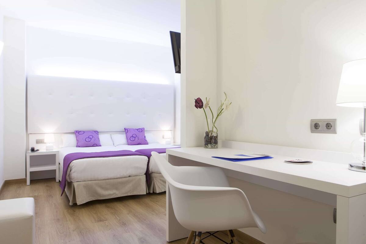 A modern hotel room with a bed, bedside table, desk, chair, and a vase with flowers.