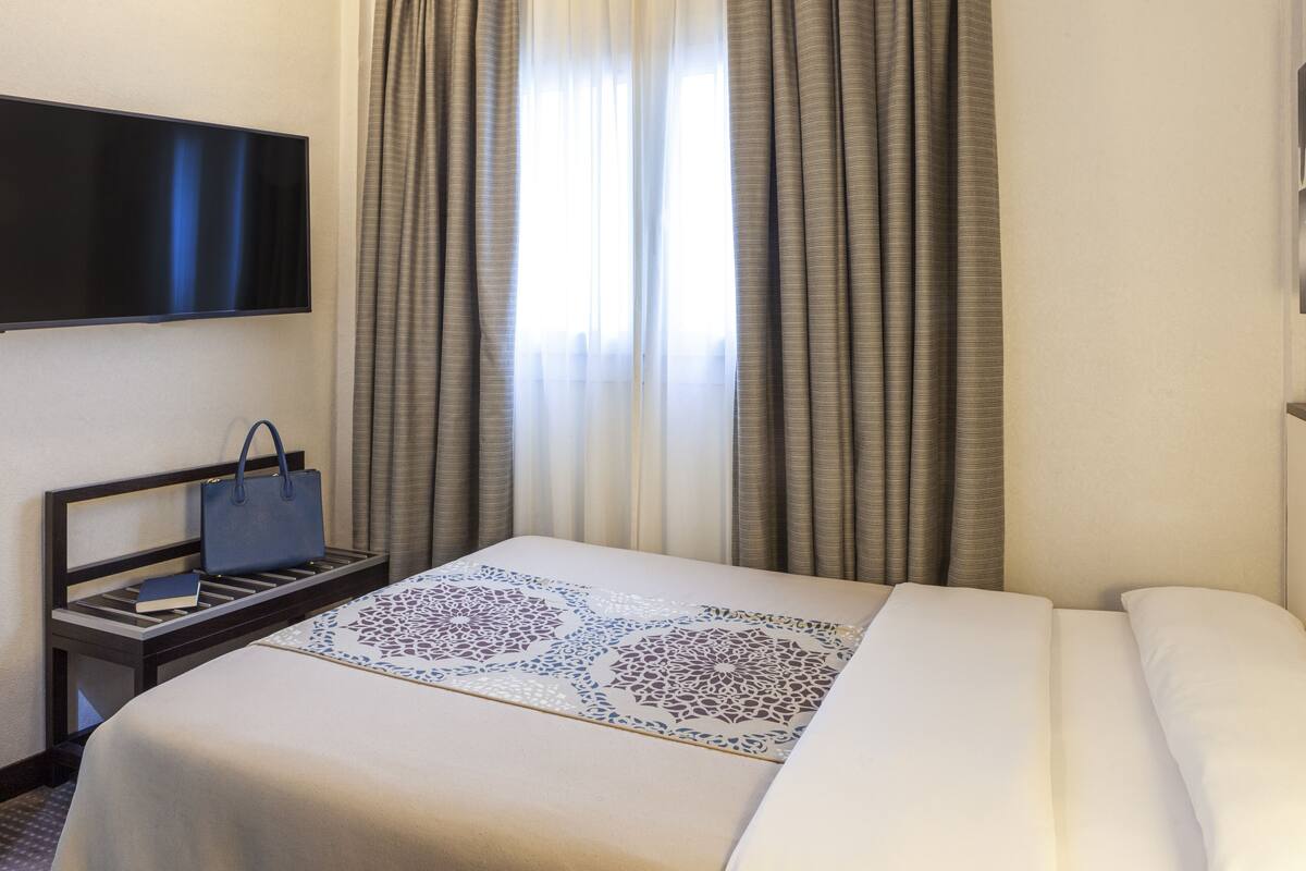 Habitación de hotel con cama, televisor montado en la pared, mesita de noche con un libro y un bolso, y ventana con cortinas.