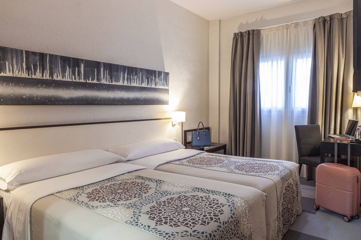Habitación de hotel con dos camas, un escritorio, una silla, una maleta y un cuadro en la pared.