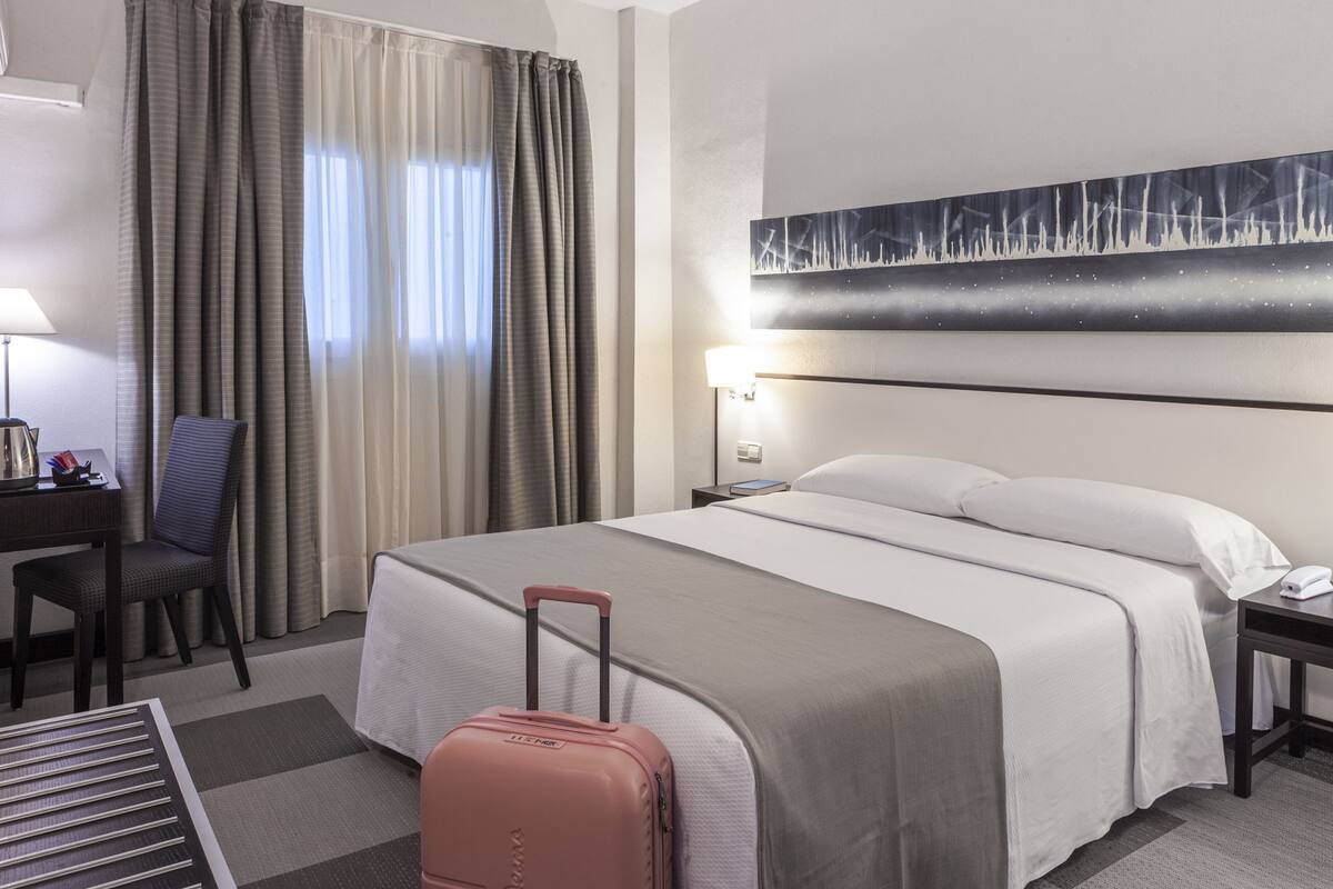 Una habitación de hotel con cama, escritorio, silla, lámpara y maleta.