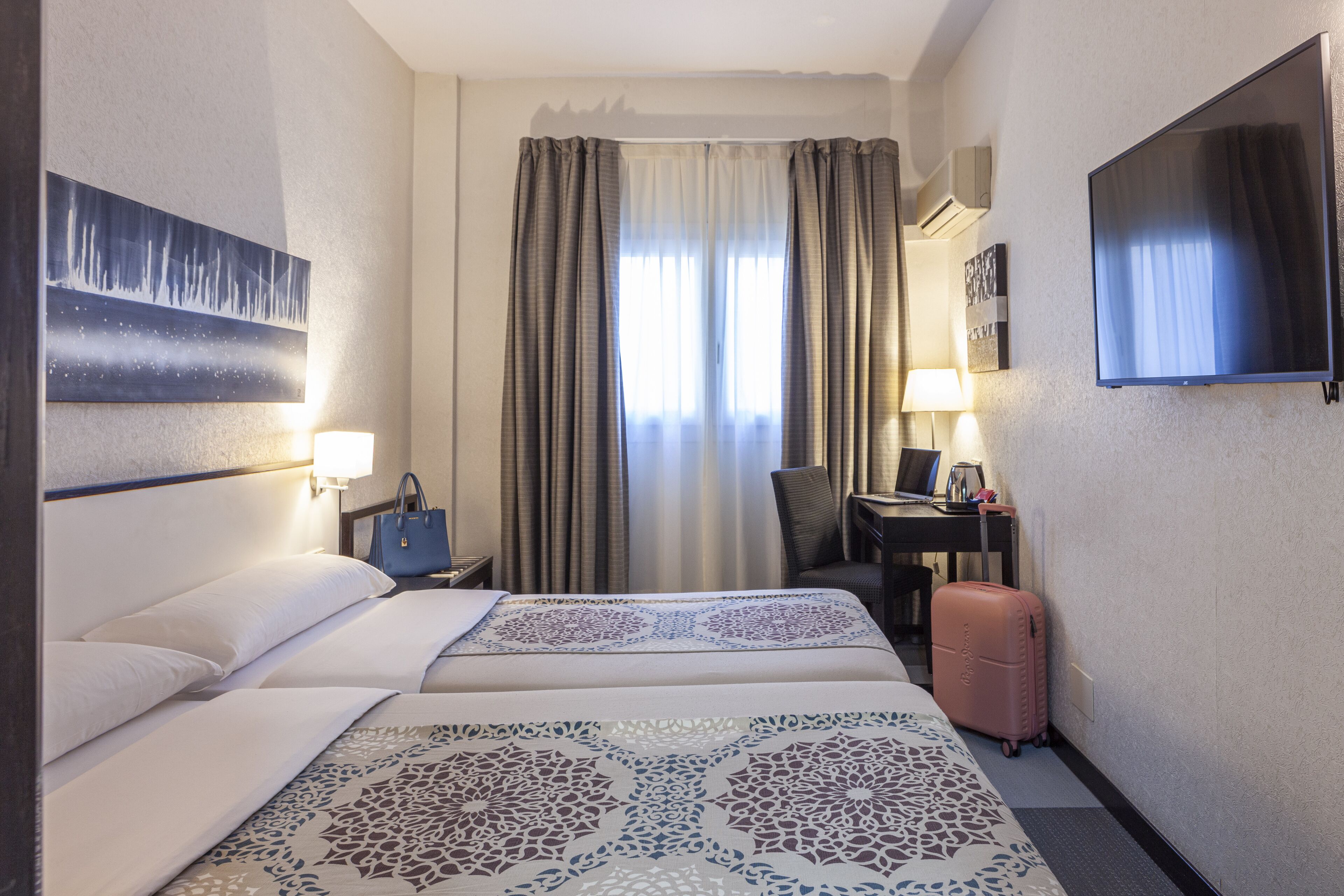 Een hotelkamer met twee bedden, een bureau met een computer, een televisie en een koffer.