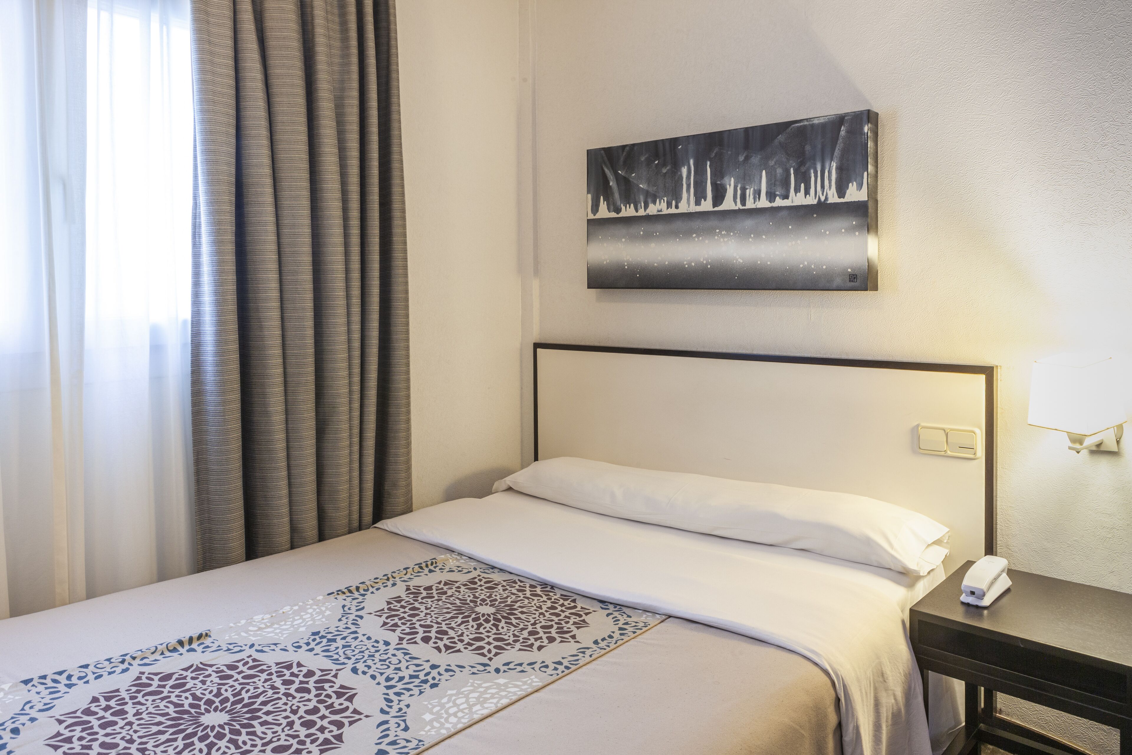 Una habitación de hotel con cama, mesita de noche, un cuadro y una ventana con cortinas.