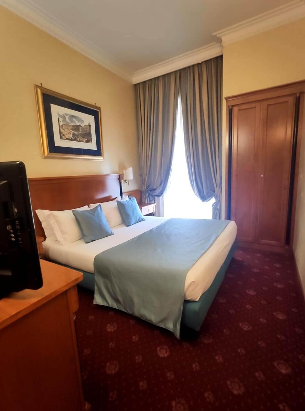 A hotel room with a bed, a desk with a computer, a framed picture on the wall, and a window with curtains.