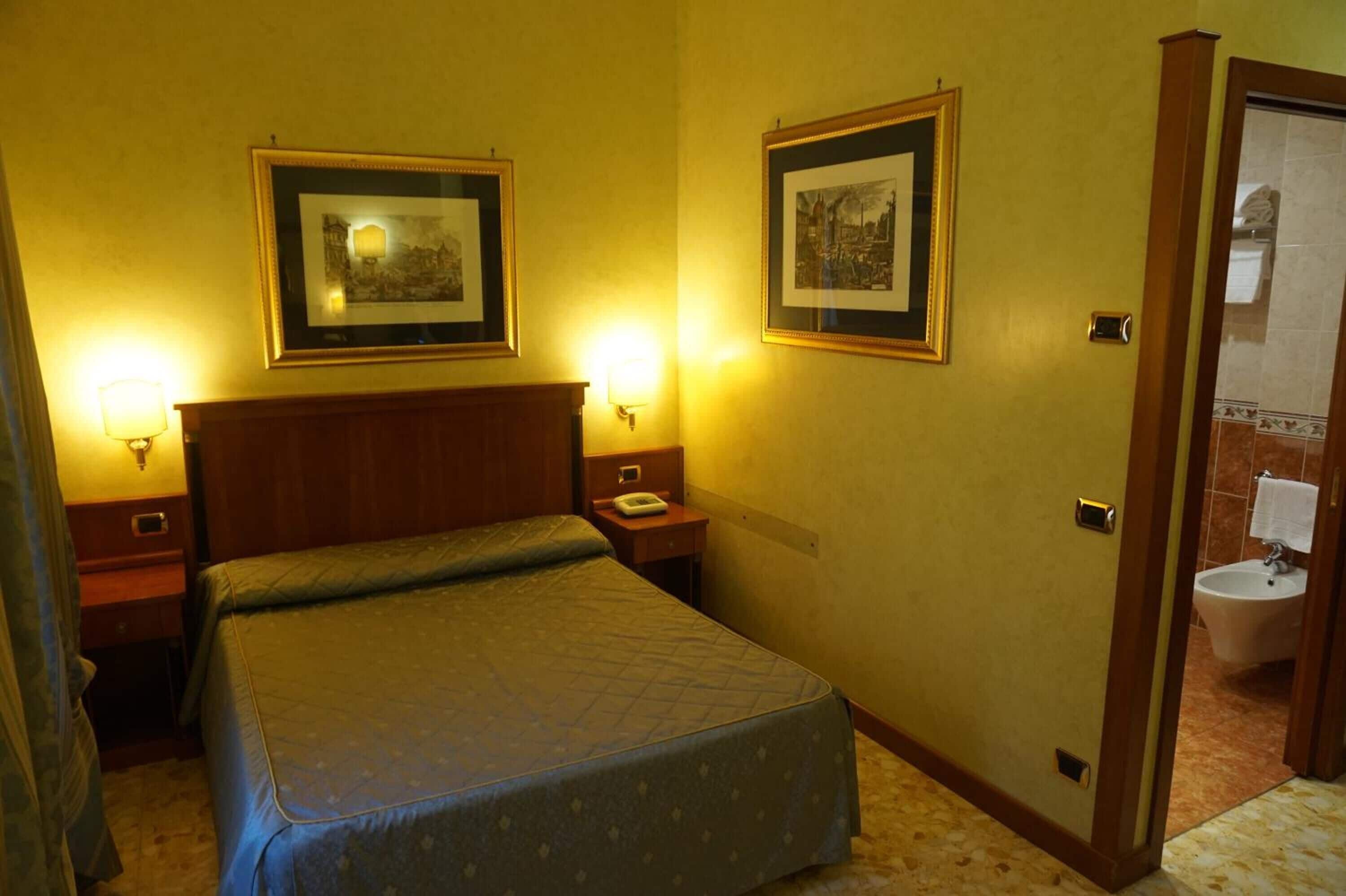 Une chambre d’hôtel avec un lit, des lampes de chevet, une table de nuit et des tableaux encadrés au mur.