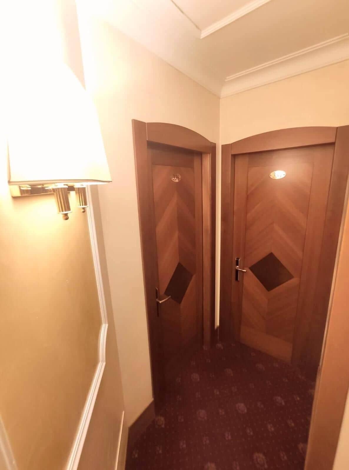 A narrow hallway with two wooden doors, each with a small window and a handle. A wall-mounted lamp is visible on the left side.