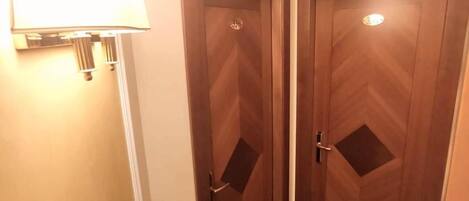 A narrow hallway with two wooden doors, each with a small window and a handle. A wall-mounted lamp is visible on the left side.