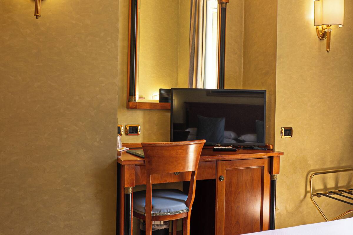 Una habitación de hotel con una cama, un escritorio con una silla, un televisor y dos lámparas fijadas a la pared.