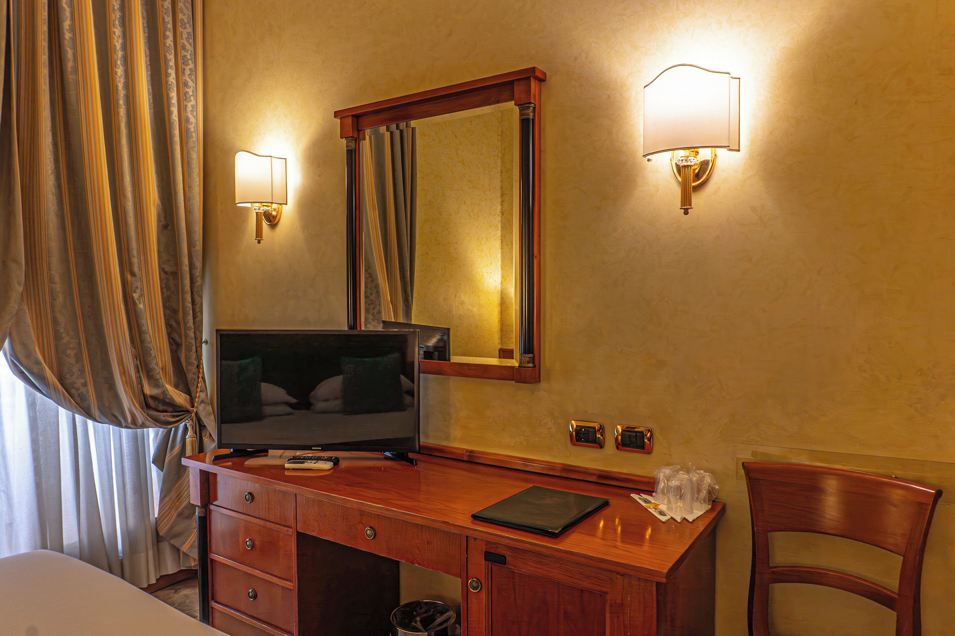 A hotel room with a wooden desk, a television, a mirror, and a lamp.