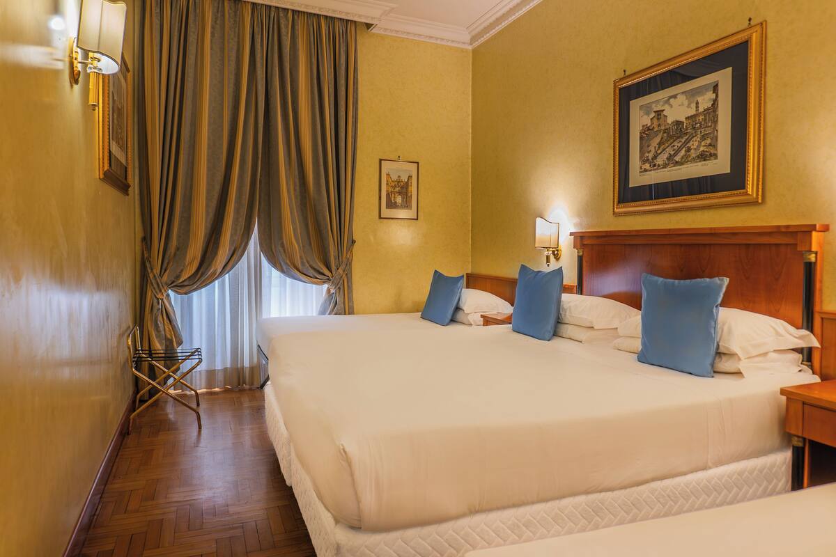 Una habitación de hotel con una cama grande, piso de madera, ventana con cortinas, mesita de noche y un cuadro enmarcado en la pared.