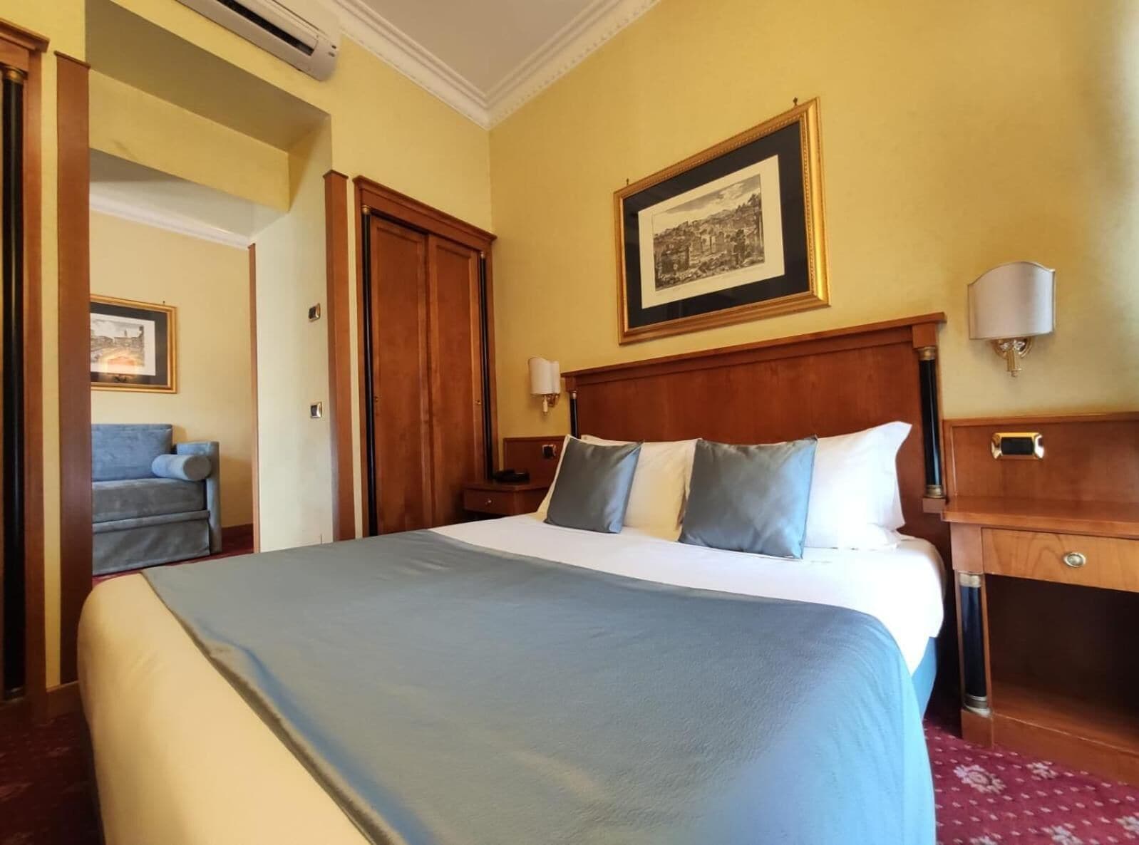 A hotel room with a large bed, a wooden headboard, a framed picture on the wall, and a nightstand with a lamp.