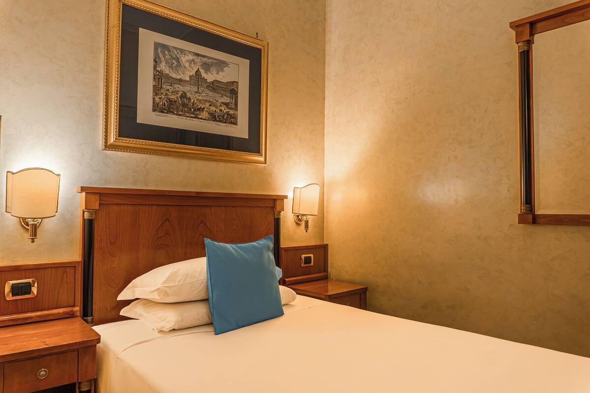 Una habitación de hotel con una cama, una mesita de noche, una lámpara de pared y un cuadro enmarcado.