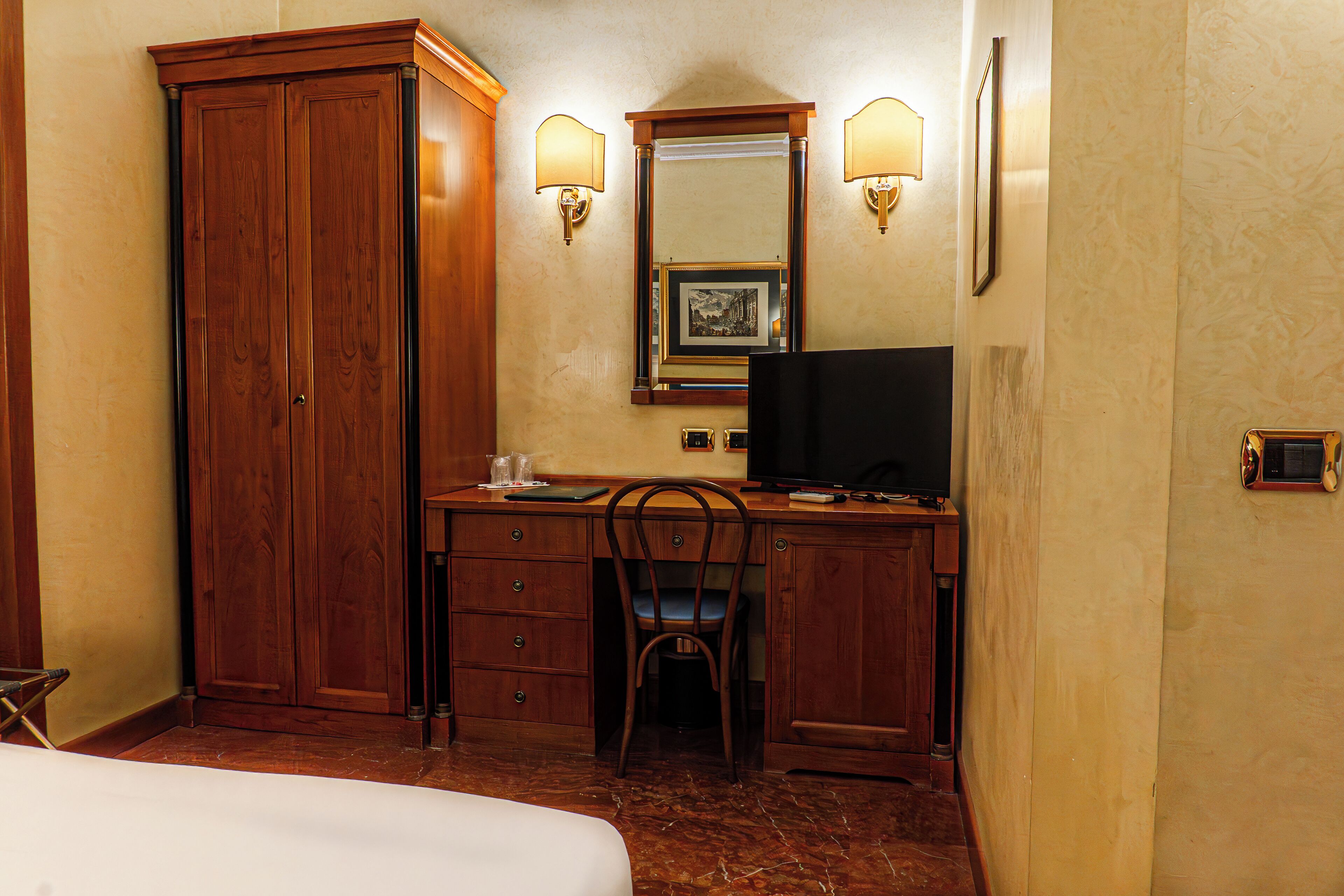 A hotel room with a wooden desk, a television, a chair, a wardrobe, and a bed.