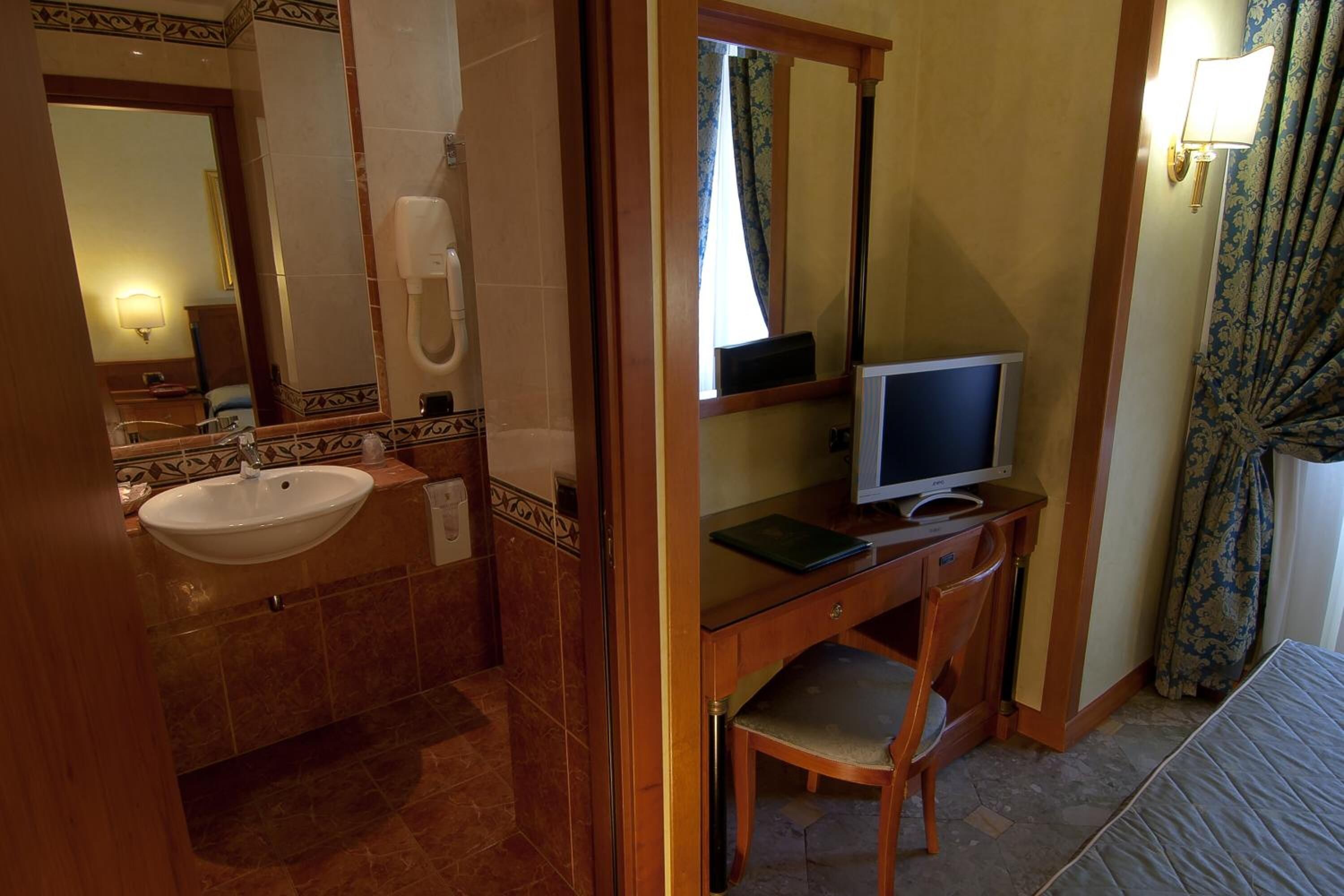A bathroom with a sink, mirror, toilet, and a desk with a computer monitor.