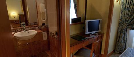A bathroom with a sink, mirror, toilet, and a desk with a computer monitor.