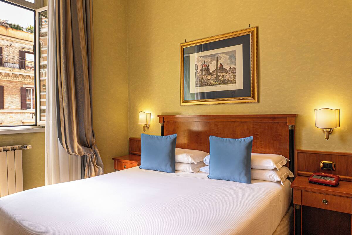 Habitación de hotel con una cama grande, dos almohadas azules, un cabecero de madera, un cuadro enmarcado en la pared y una mesita de noche con una lámpara y un teléfono.