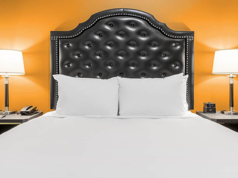 Habitación de hotel con cabecera capitonada, ropa de cama blanca, dos mesitas de noche con lámparas y un reloj.