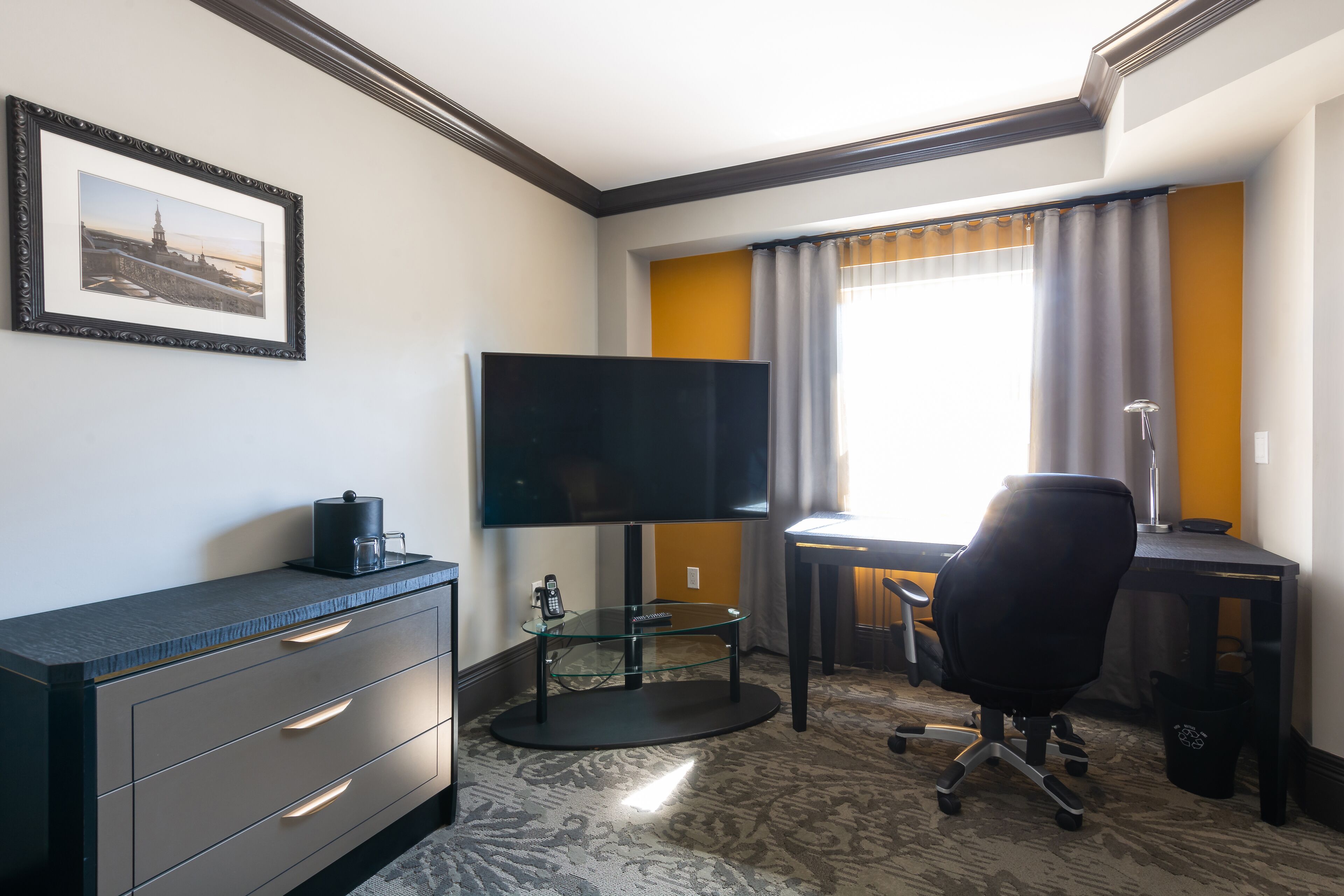 Habitación de hotel con televisión de pantalla plana, escritorio con silla, mesa de centro de vidrio y un cuadro en la pared.