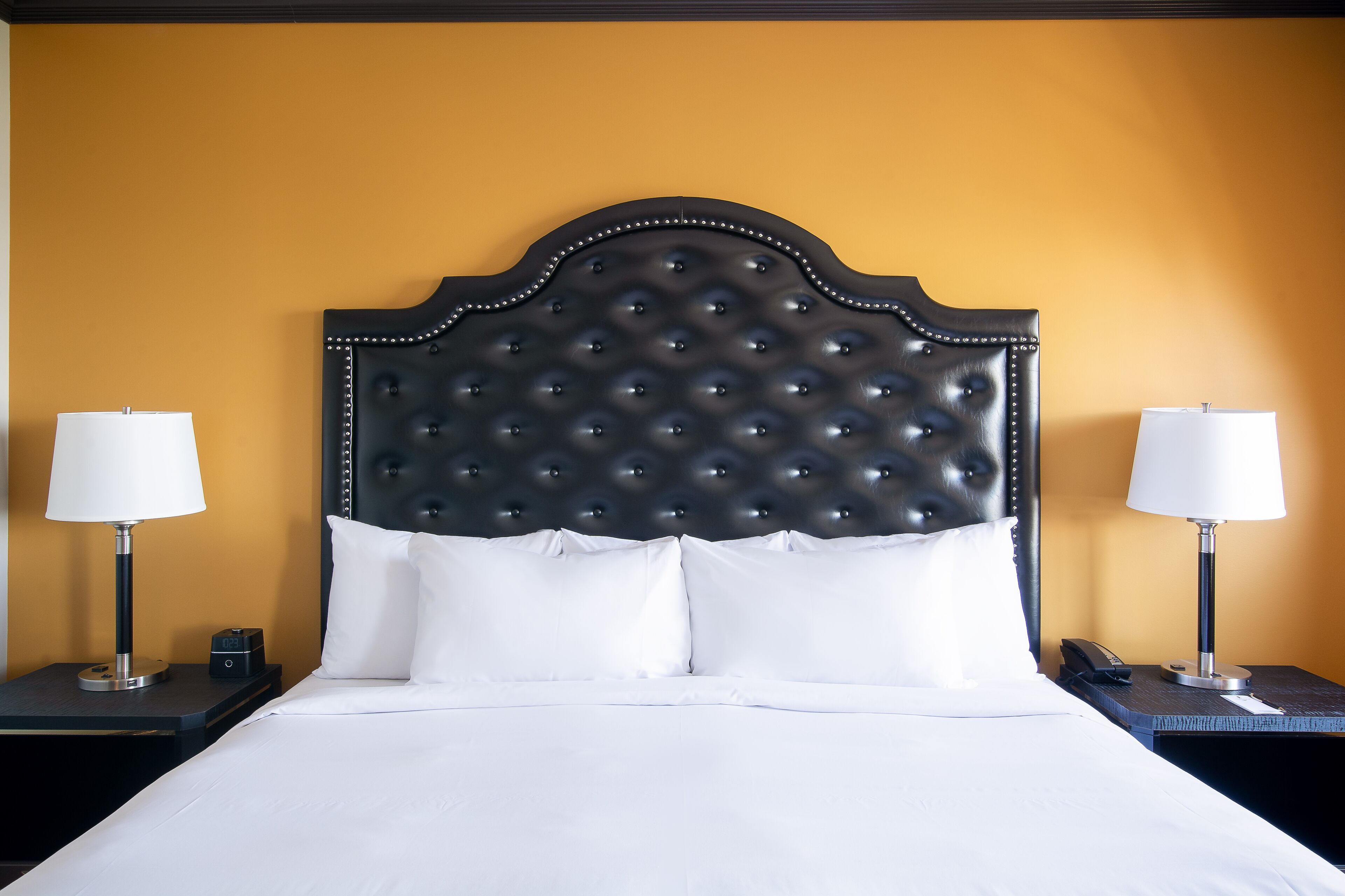 Una cama bien tendida con un cabecero capitonado, dos mesitas de noche con lámparas y una pared lisa en un tono amarillo cálido.