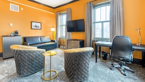 Quarto de hotel com sofá, dois pufes, escrivaninha, cadeira, televisão e uma mesa pequena.