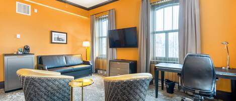 Une chambre d’hôtel comprenant un canapé, deux poufs, un bureau, une chaise, une télévision et une petite table.