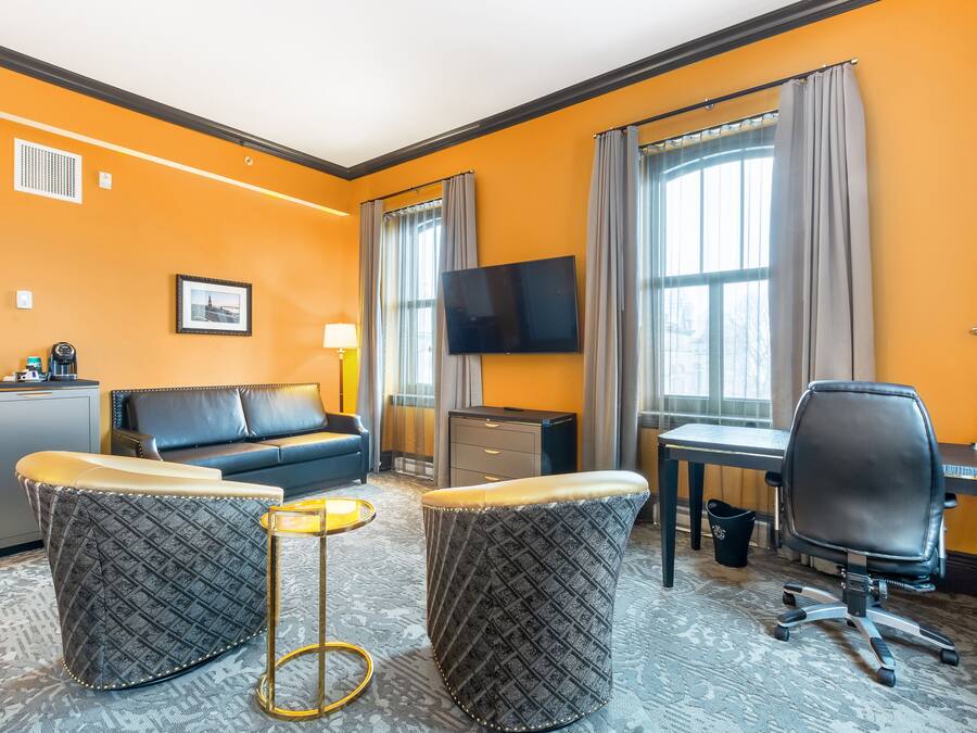 Habitación de hotel con sofá, dos otomanas, escritorio, silla, televisor y una mesita.