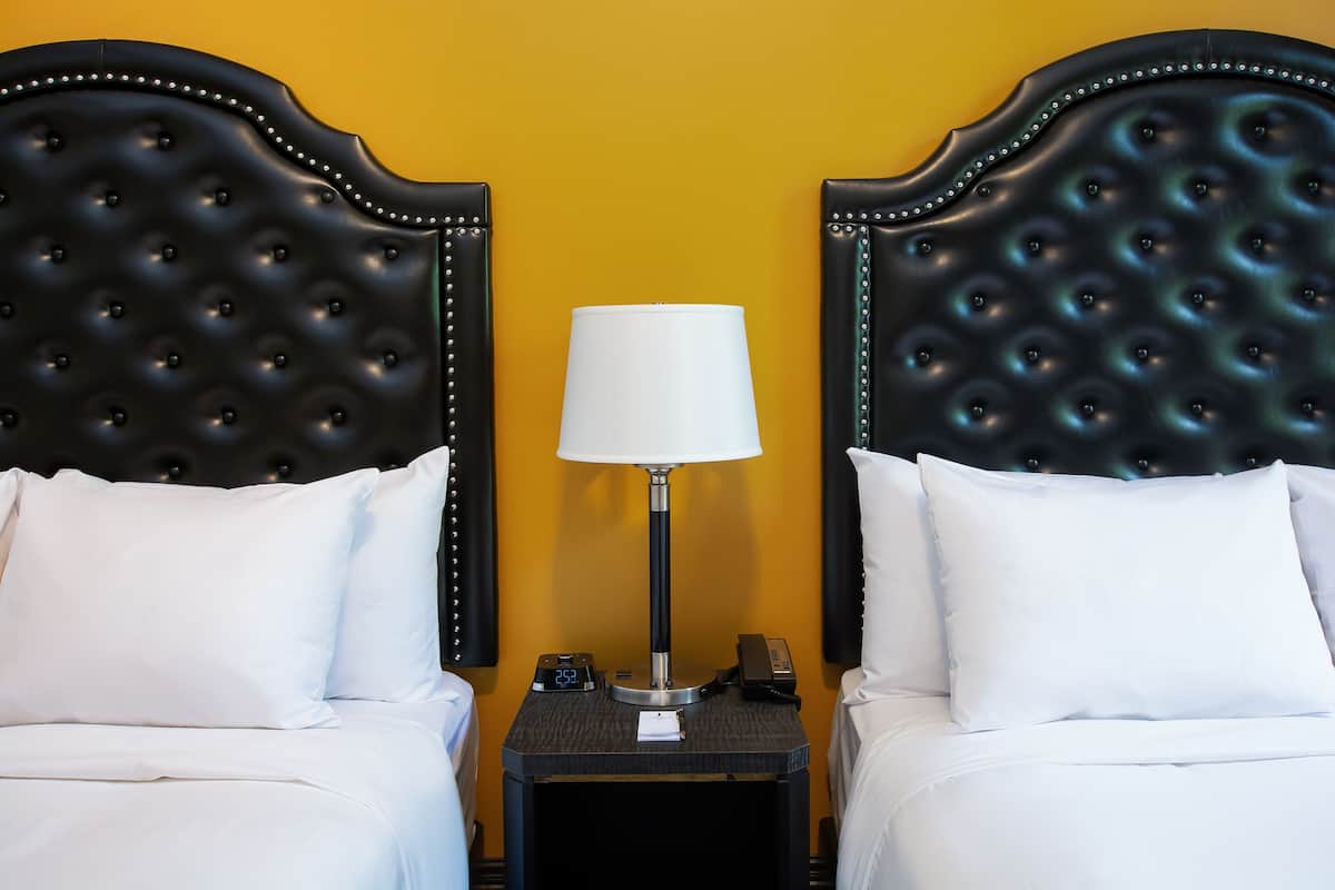 A hotel room with two beds, a nightstand with a lamp, a clock, and a phone.