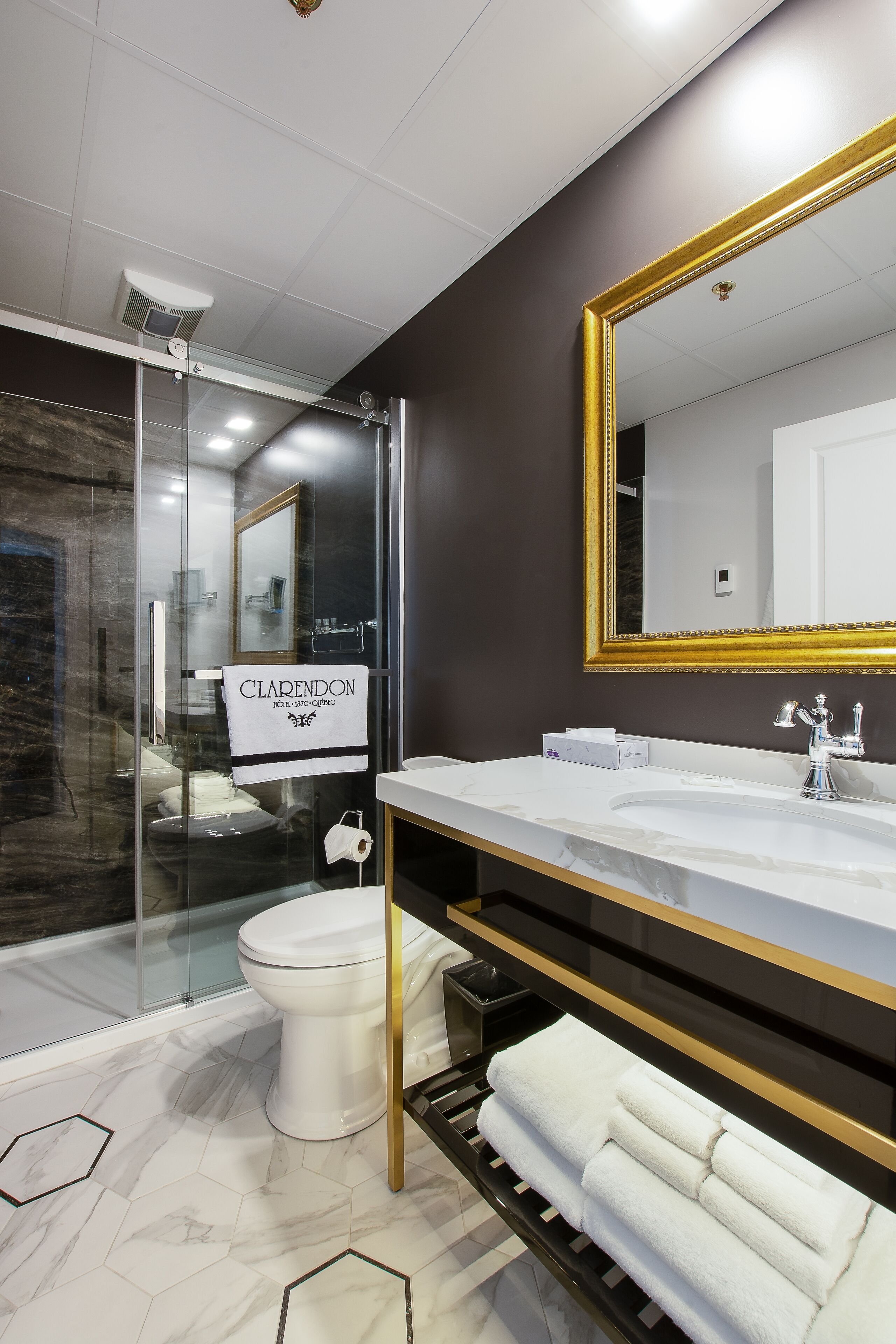 A modern bathroom with a glass shower enclosure, a white toilet, a large mirror with a gold frame, and a marble-tiled floor.