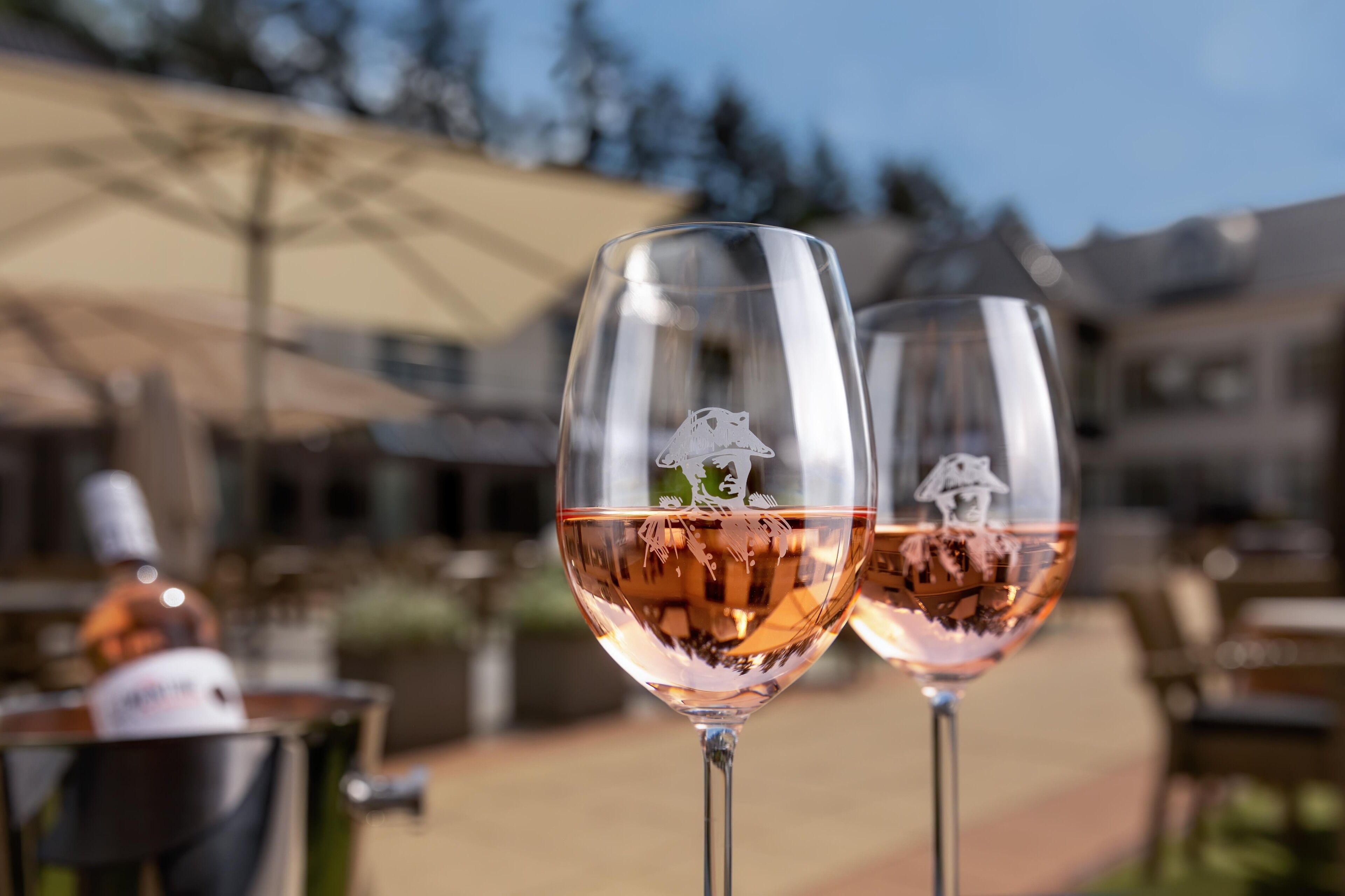 Two wine glasses with a reflection of a person and a dog, placed outdoors with a blurred background of tables and umbrellas.