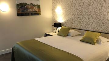 Een net opgemaakt bed met een groene deken en twee kussens, een nachtkastje met een lamp, en een ingelijste foto aan de muur.