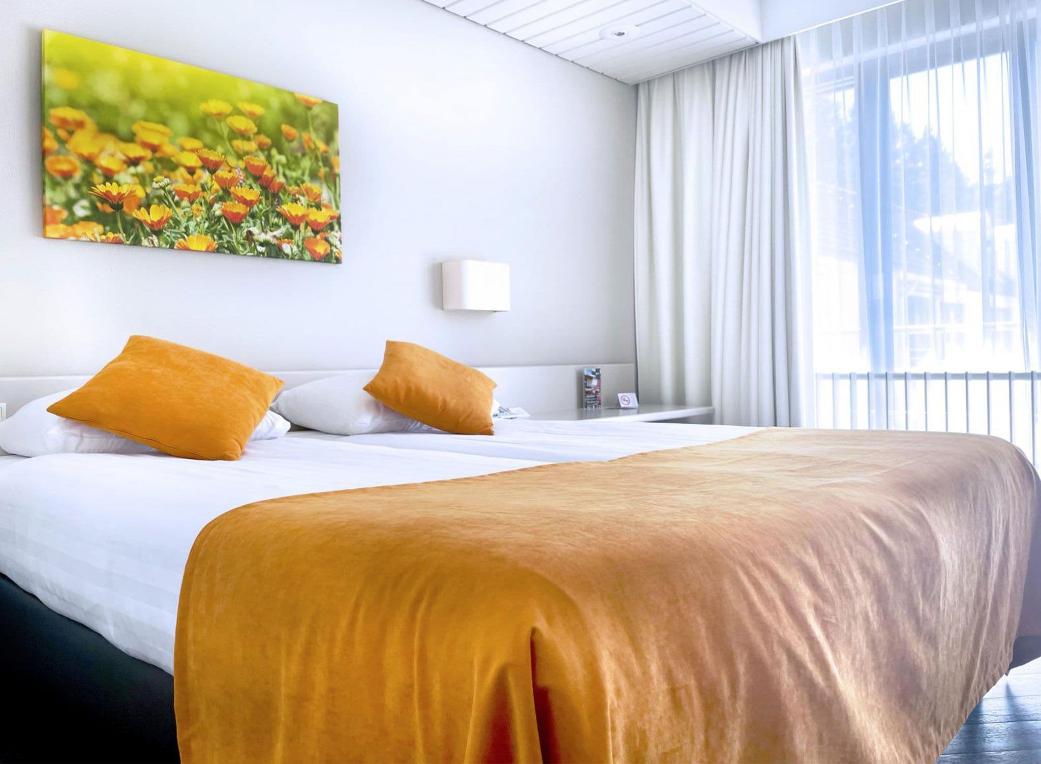 Une chambre d’hôtel avec un grand lit, deux oreillers, un tableau de fleurs, une fenêtre avec des rideaux et une table de chevet.