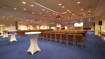 Un salón de conferencias amplio con filas de sillas y mesas, una pantalla de proyector y suelo alfombrado.