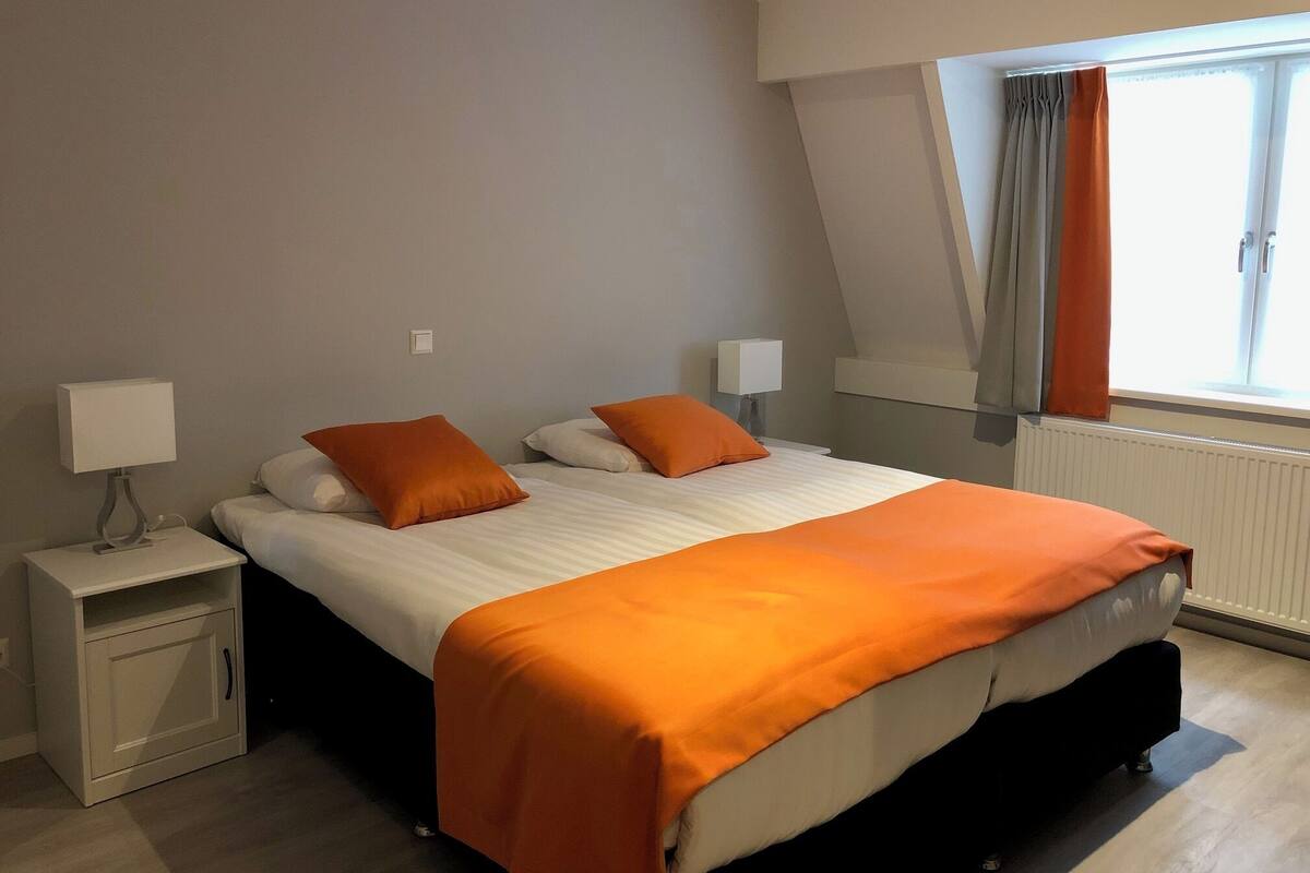 A bedroom with a bed, two bedside tables, a window with curtains, and a radiator.
