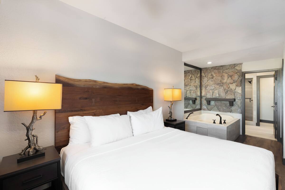 Una habitación de hotel moderna con una cama grande, dos lámparas de noche, un cabecero de madera, una pared de piedra, un baño con bañera y zona de ducha.