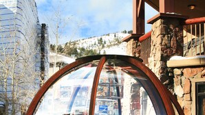 A wooden gazebo with a glass roof, surrounded by snow, next to a stone building with a wooden balcony.