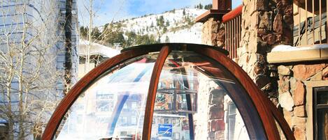 Un pabellón de madera con techo de cristal, rodeado de nieve, junto a un edificio de piedra con balcón de madera.