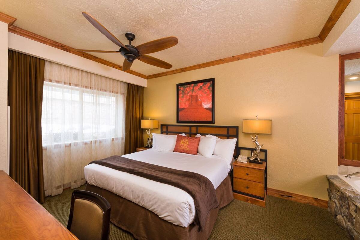 Habitación de hotel con cama, escritorio, silla, ventilador de techo, mesita de noche con lámpara, un cuadro en la pared y una pared decorativa de piedra.