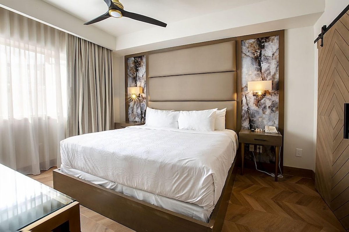 A hotel room with a large bed, a desk, a ceiling fan, and two wall-mounted lamps.