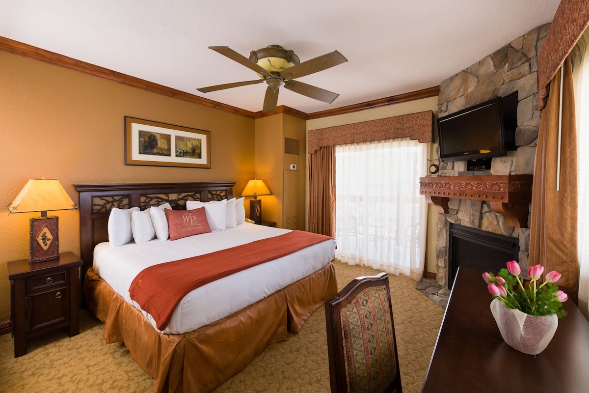 Habitación de hotel con una cama, ventilador de techo, televisión instalada en una pared de piedra, chimenea y un florero con flores sobre una mesa.
