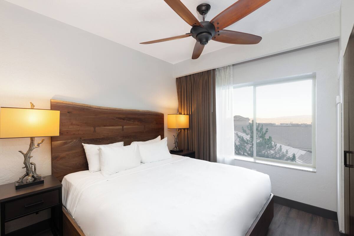 Habitación de hotel con una cama grande, dos lámparas de noche, un ventilador de techo, una mesita de noche, un ventanal con vistas y un cabecero de madera.