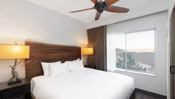 Habitación de hotel con una cama grande, dos lámparas de noche, un ventilador de techo, una mesita de noche, un ventanal con vistas y un cabecero de madera.