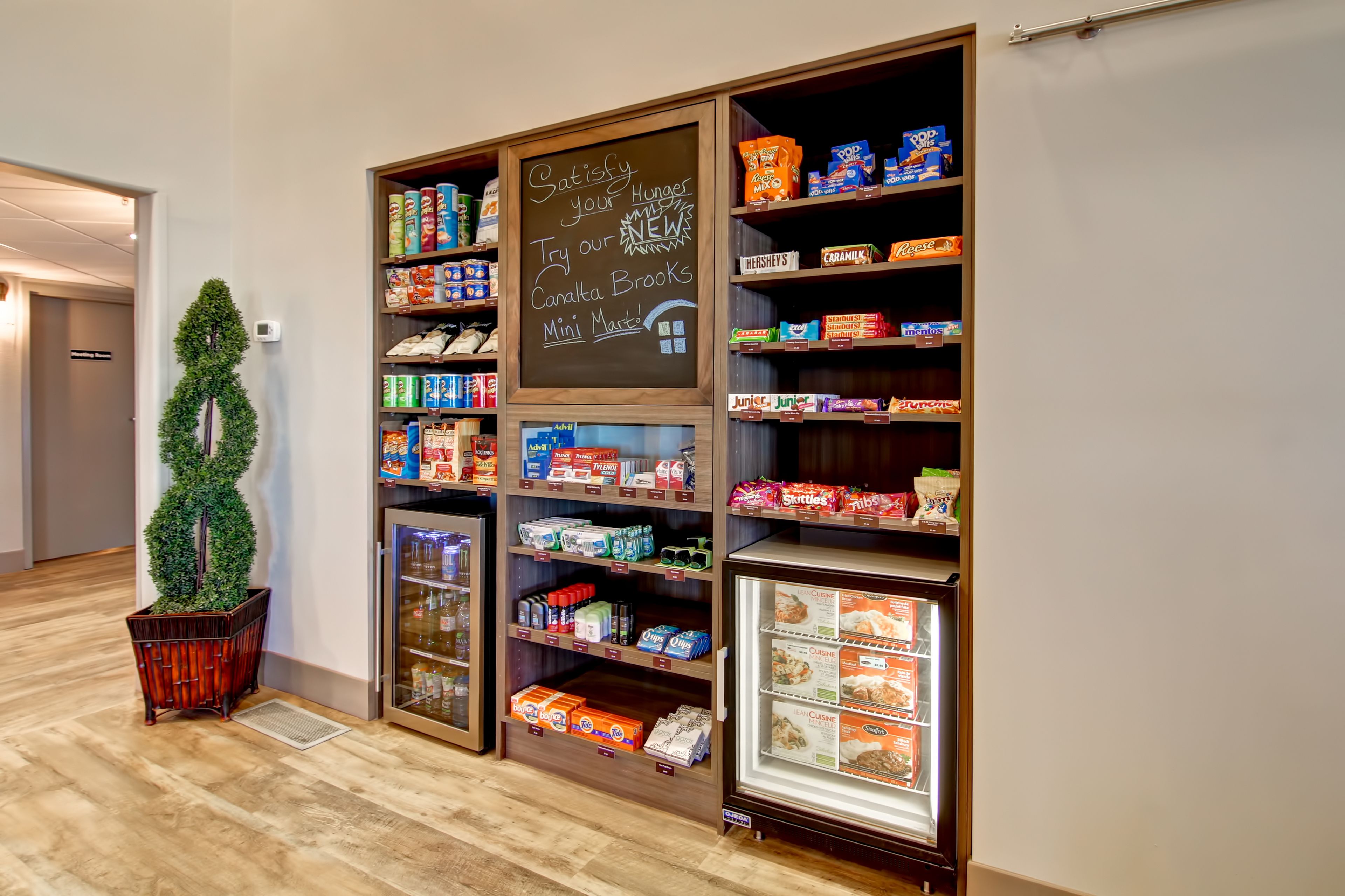 A mini market with shelves stocked with snacks and beverages, a chalkboard sign, and a decorative tree.