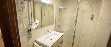 Une salle de bain avec un mur en marbre, un lavabo blanc, un miroir, un sèche-cheveux et une douche à parois de verre.