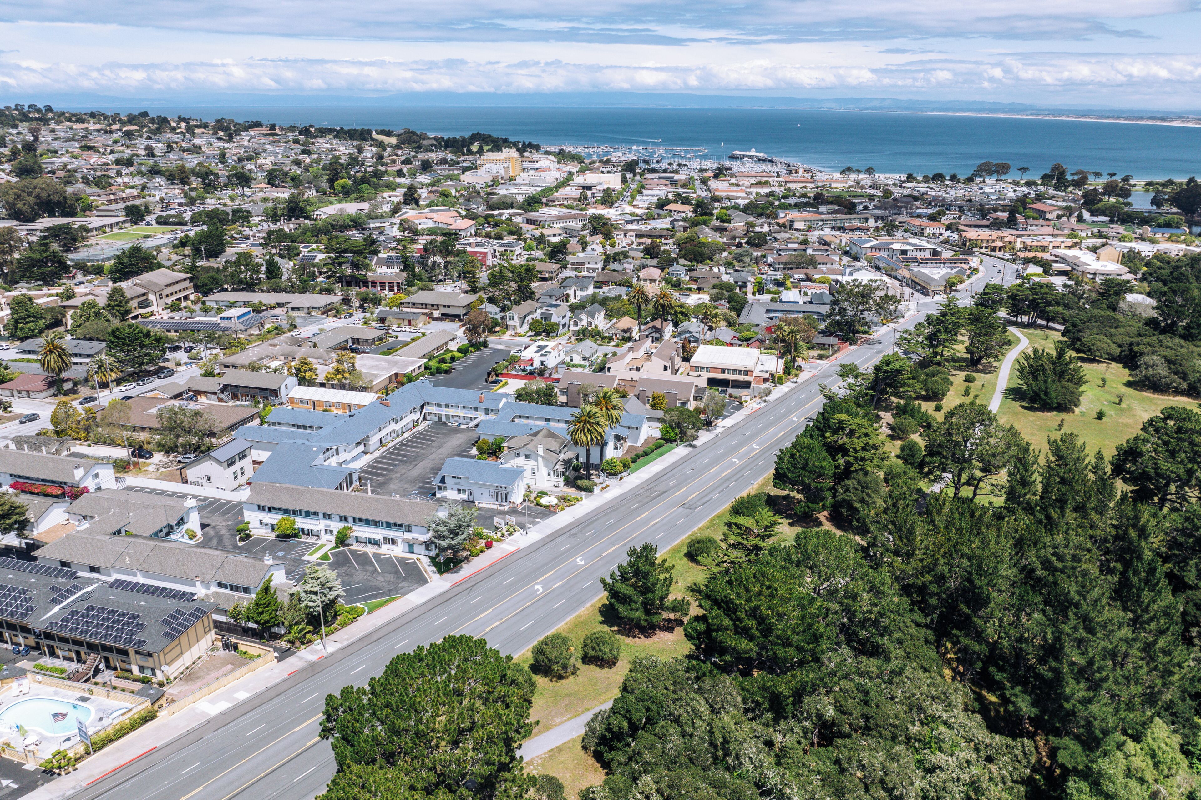 An aerial view of a residential area with houses, a main road, and a coastline in the background.