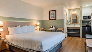 A hotel room with a bed, bedside tables, a desk, a chair, a small table, a microwave, a refrigerator, and a mirror.